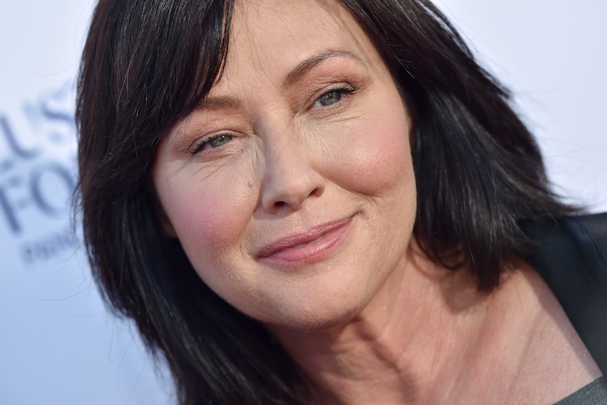 A close up of Shannen Doherty's face, smiling, long dark hair, greyish blue eyes