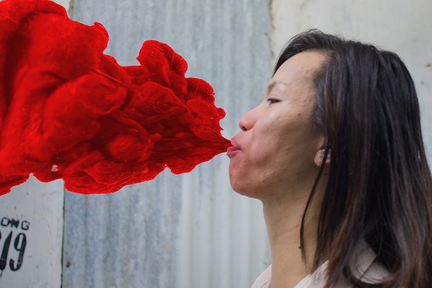 Colour still of a woman blowing out a graphic of red substance from 2014 short film The Living Need Light, the Dead Need Music.