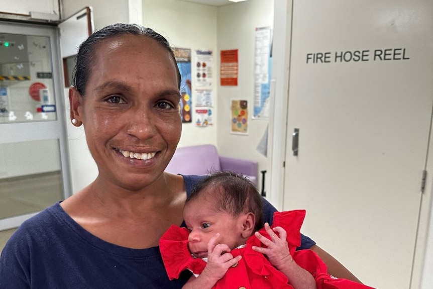 New mother smiling in hospital while holding her newborn daughter who is wearing a red frilly dress.