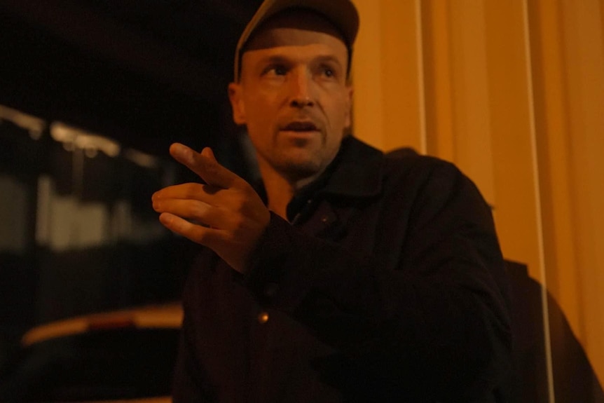 A man wearing a cap pointing in a dimly lit street. 