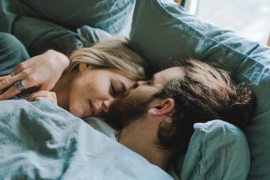 Man and woman cuddling in bed, with man kissing partner on her forehead for a story about the rising unpopularity of the pill.