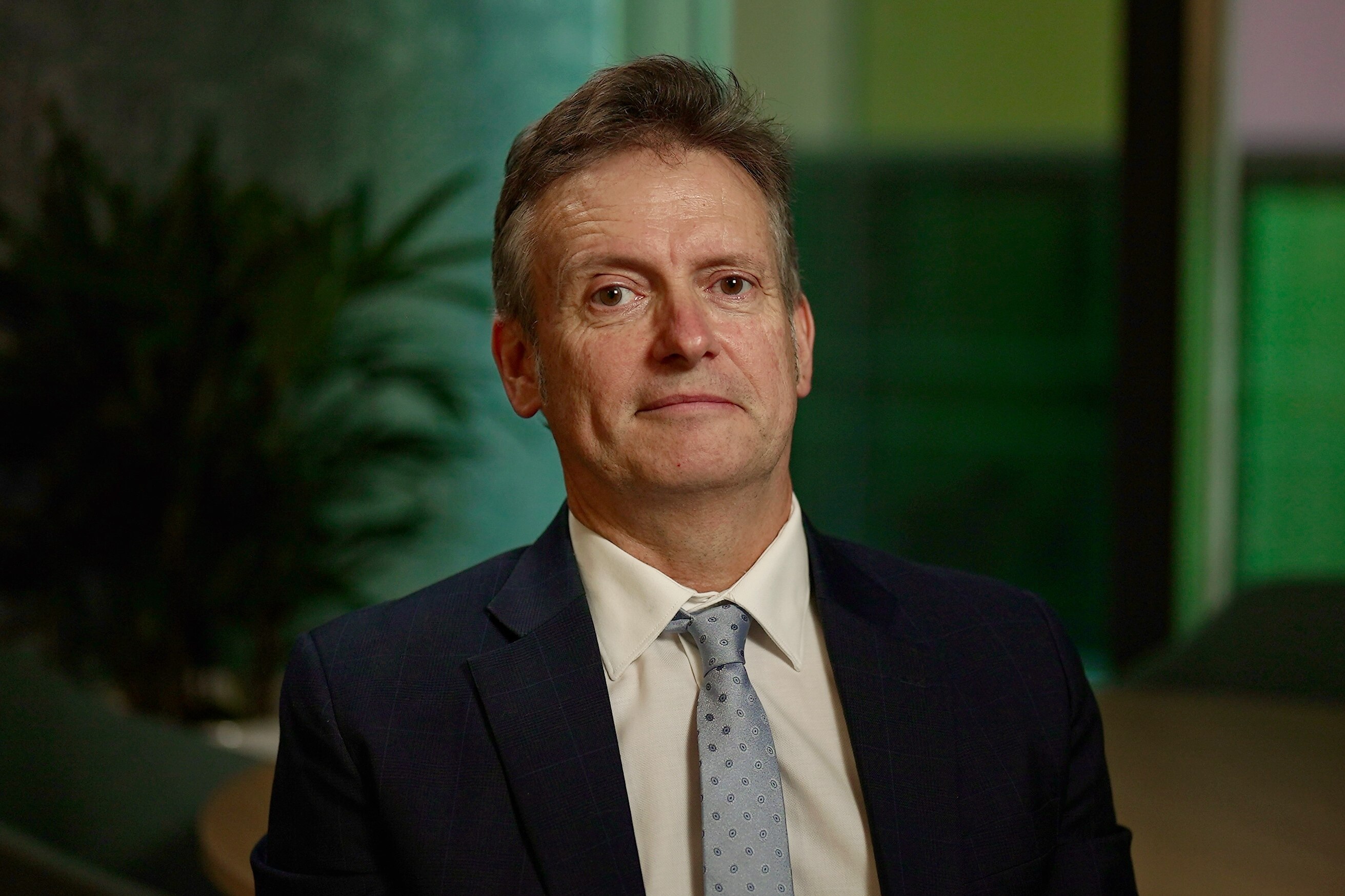 A portrait of Scott McDougall wearing a suit jacket, white shirt and light blue tie in front of a green background.