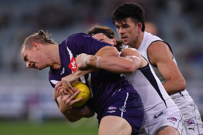 A Fremantle AFL player holds the ball as he is tackled around his arms by a Carlton opponent.