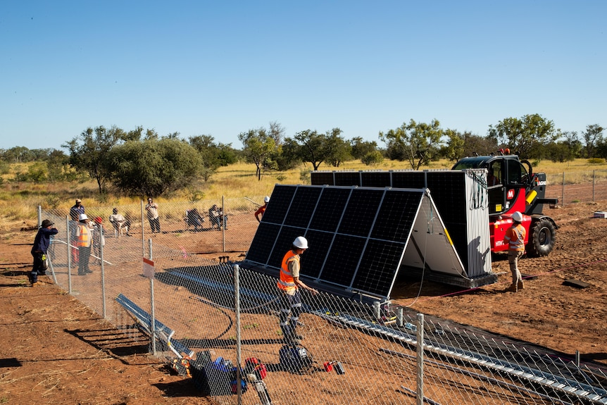 a photo showing a group of people working on a solar panels with others standing behind a fence and looking on.