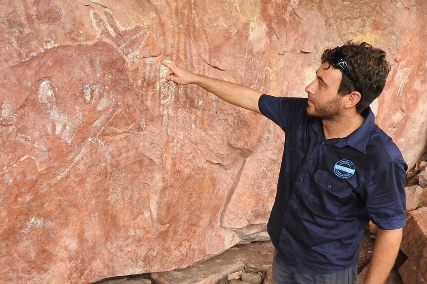 A dark-haired man in a branded work shirt points at an engraving on a sandstone wall.
