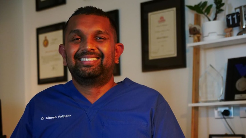 A man wearing blue medical scrubs smiles at the camera. Certificates are framed on the wall behind him.