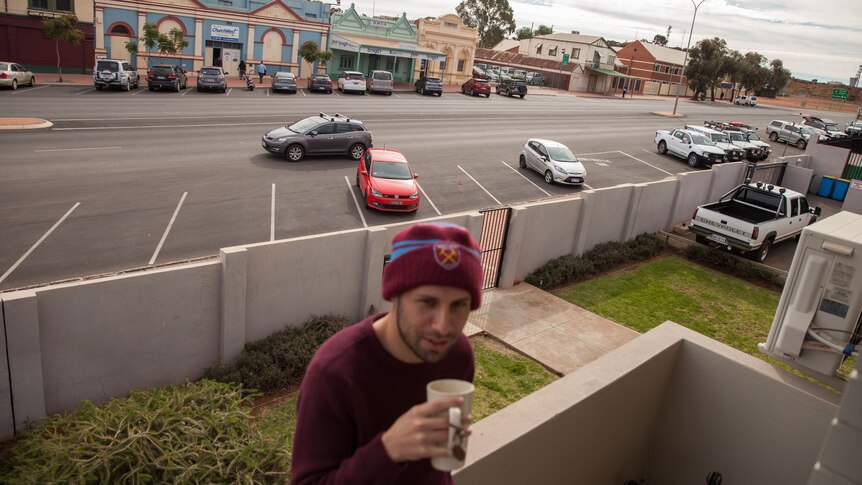 A young man in beanie drinks coffee on a balcony, the street of a country town below.
