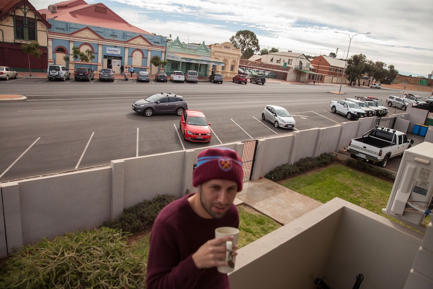 A young man in beanie drinks coffee on a balcony, the street of a country town below.