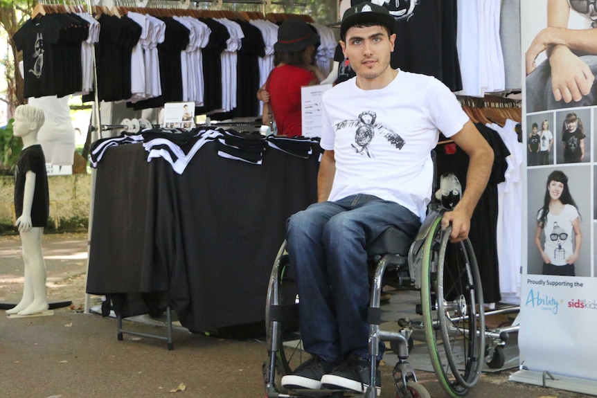 A man sits in his wheelchair outside a t-shirt stall with racks of white and black shirts.