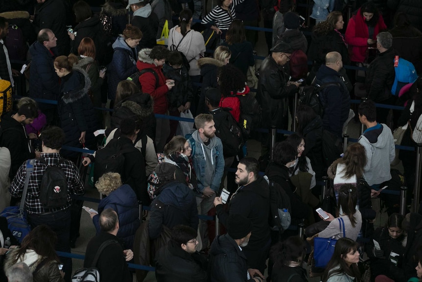 One man in a crowd looks to the distance as everyone around him looks down or away from the camera while waiting at JFK airport.