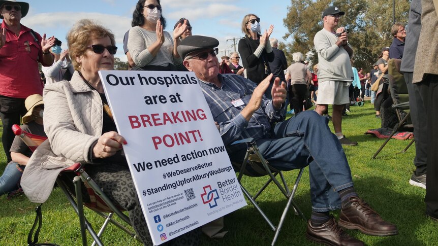 A man and woman sit on picnic seats at a rally and woman holds up a sign warning about hospitals being under stress. 