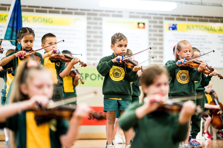 Primary school students in green and yellow uniforms play stringed instruments on a school hall stage.