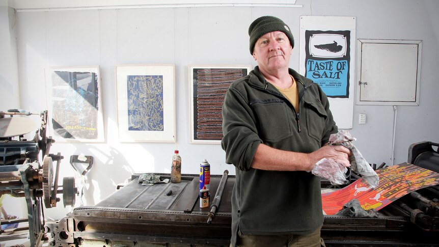 Printer Tom Goulder stands in his workshop in front of a printing press, cleaning his hands with a cloth.