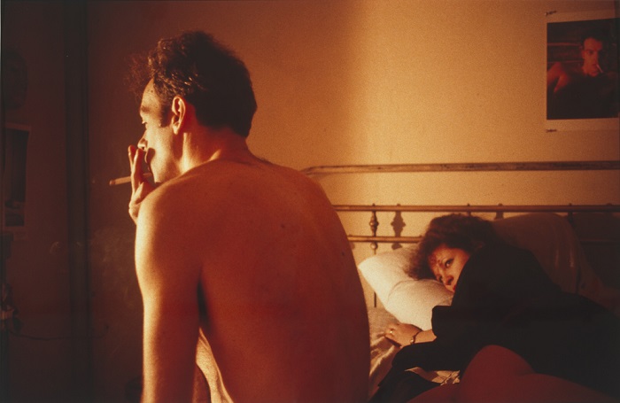 A man sits on a bed smoking while a woman lies beside him.