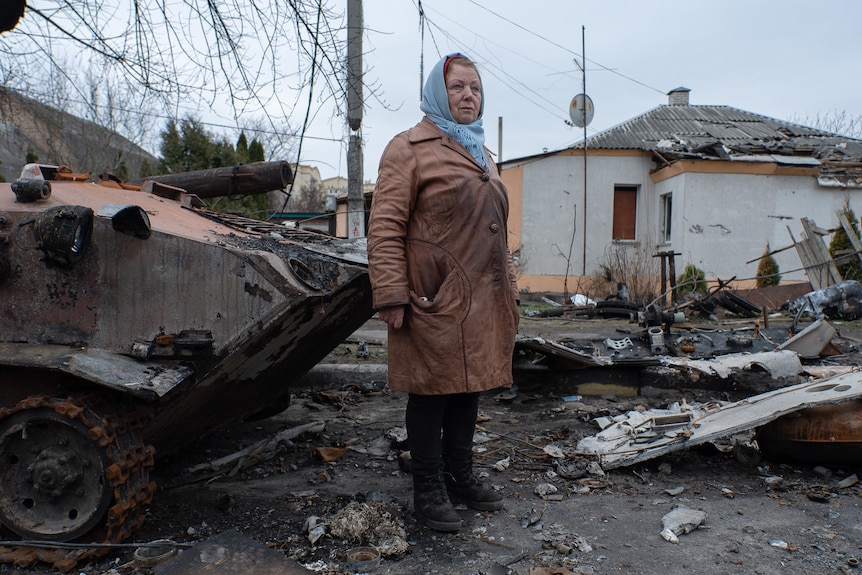 A woman wearing a blue scarf around her head and brown coat stands in a street surrounded by rubble.