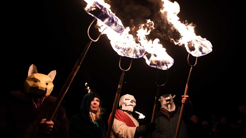 People wearing animal masks hold pitchforks with flaming sods of turf at the end.