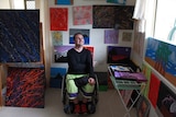 David Conway in his home surrounded by colourful paintings. Looking out the window, he has a big grin on his face.