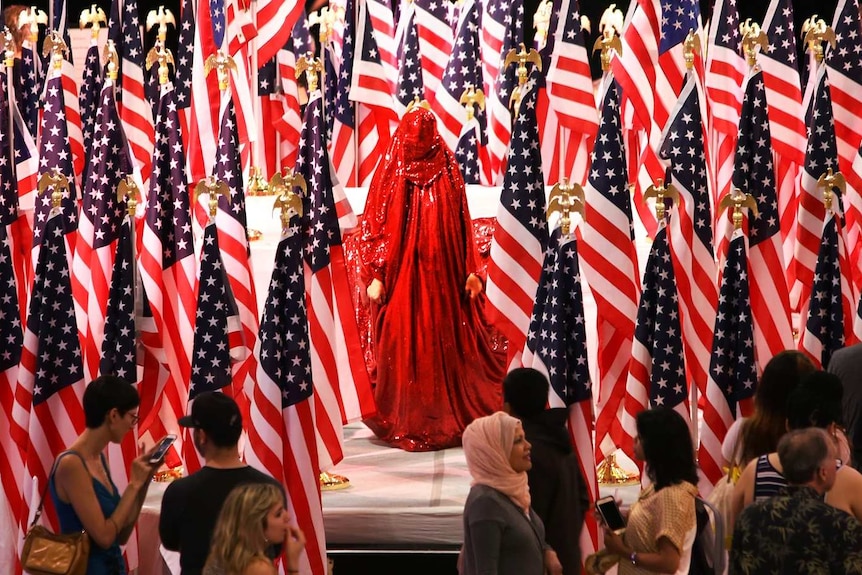 A woman in a red sequinned body covering stands in a forest of american flags, an audience watching on