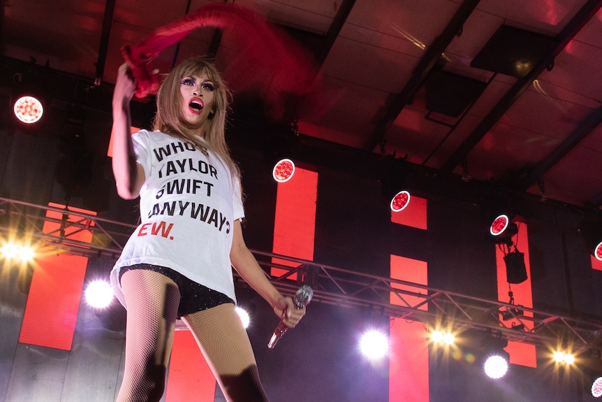 A drag queen wearing a white T-shirt that says 'Who's Taylor Swift Anyway? Ew' onstage holding a microphone and red scarf.