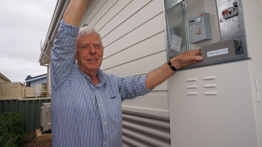 Des Clarke stands outdoors next to an open power box on the wall of his house.