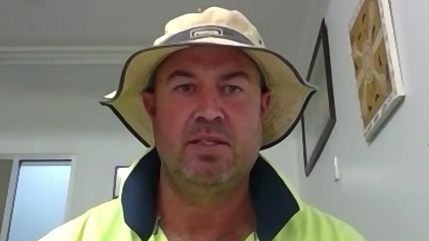 A man in a hat and a hi-vis top sits at a desk in a room on a video call.