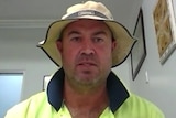 A man in a hat and a hi-vis top sits at a desk in a room on a video call.