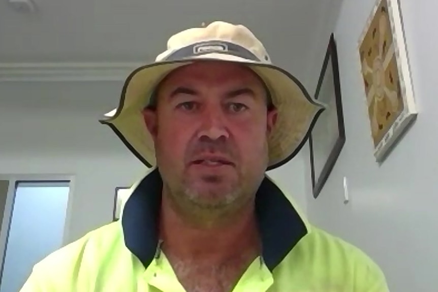 A man in a hat and a hi-vis top sits at a desk in a room on a video call.