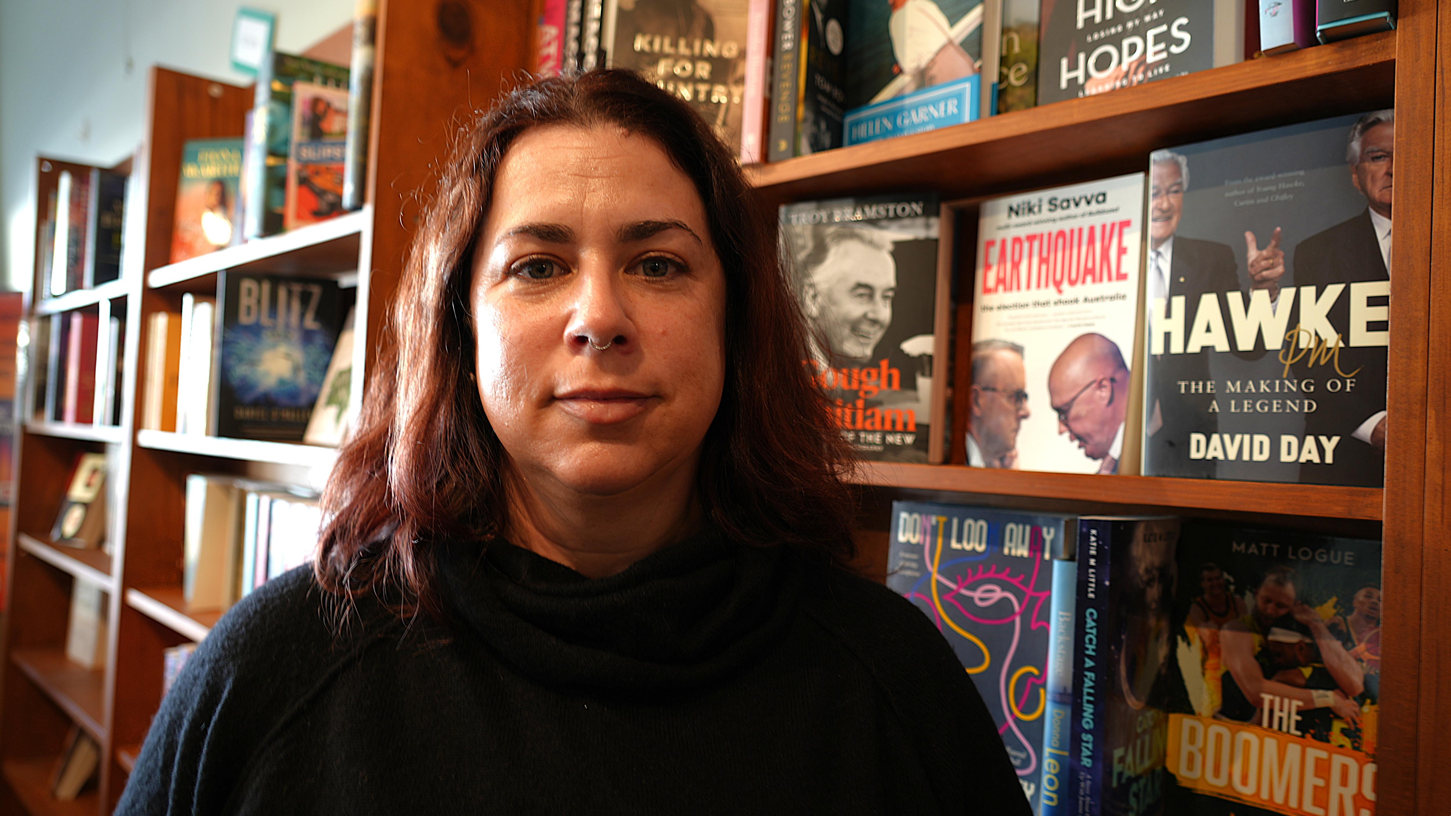 A woman in a dark jumper stands against a book case.
