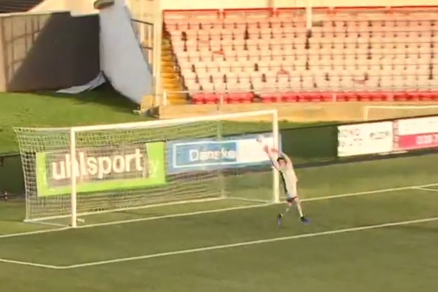 A man wearing green reaches up in the air to catch the ball in front of a soccer goal