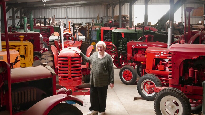 A woman stands in a large shed surrounded by vintage tractors