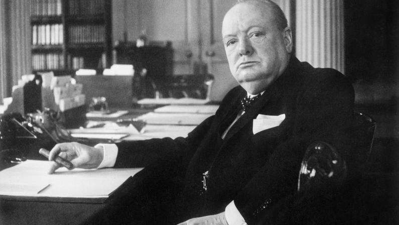 Winston Churchill is seated at his desk, looking at the camera and holding a cigar.