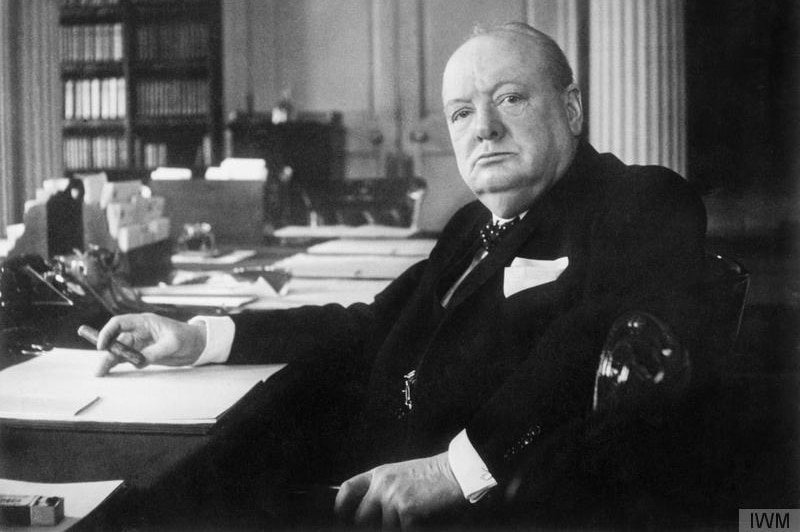 Winston Churchill seated at his desk looking at camera and holding cigar