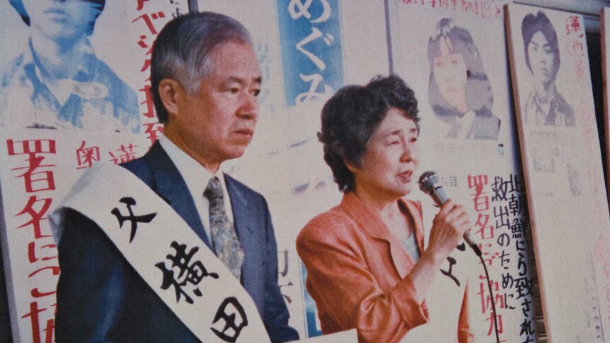 An elderly Japanese man holds up a photo of a young girl while an elderly woman speaks into a microphone