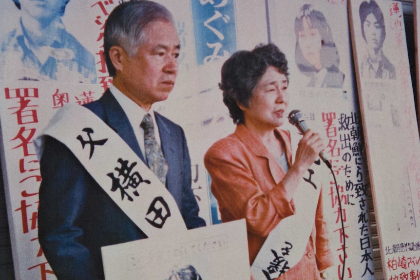 An elderly Japanese man holds up a photo of a young girl while an elderly woman speaks into a microphone