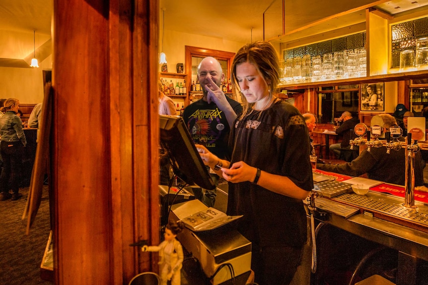 A woman stands at a computer in a busy pub putting in an order with a man behind her.