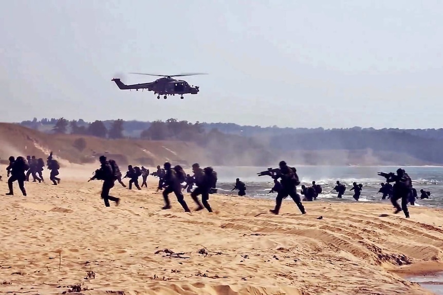 A group of soldiers storm a beach with guns raised and a military helicopter flying overhead.