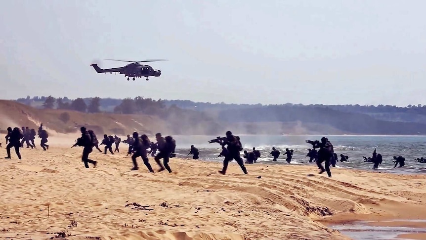 A group of soldiers storm a beach with guns raised and a military helicopter flying overhead.