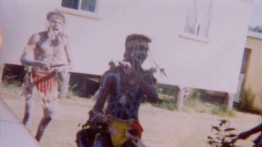 An old photograph of a young Aboriginal dancer dancing with his troupe on grass outside a building.