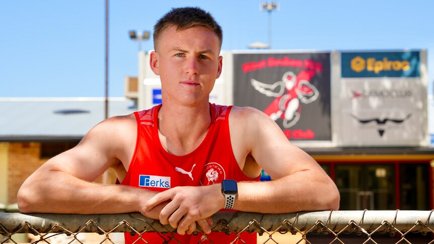 A man teenager wearing a red singlet leaning up against a fence outside the West Broken Hill Football Club