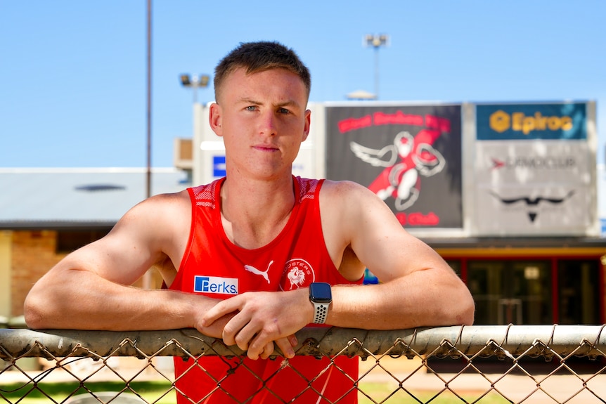 A man teenager wearing a red singlet leaning up against a fence outside the West Broken Hill Football Club