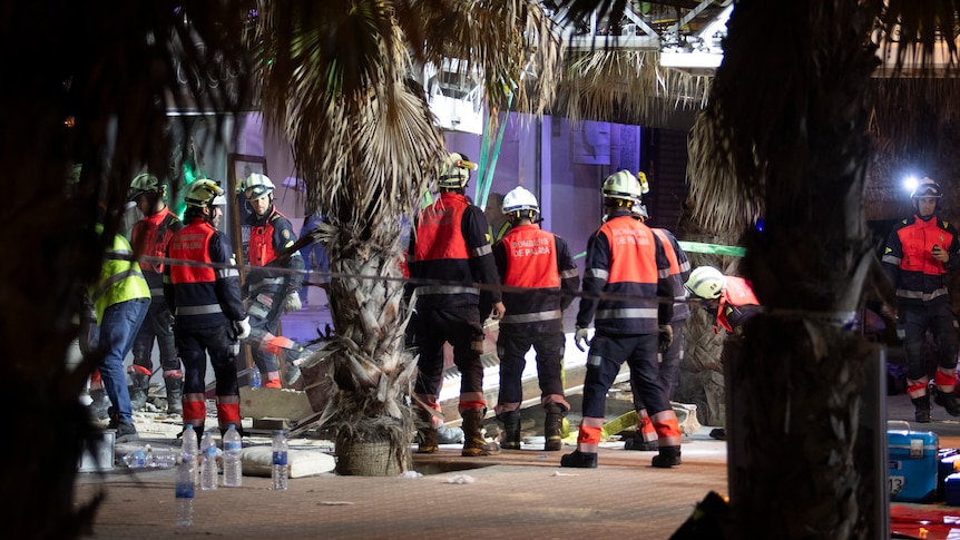 People wearing hard hats and high vis stand around a collapsed building. 