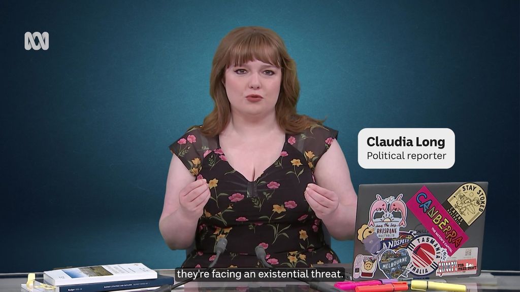 Reporter with long hair wearing floral blouse, seated at desk with a laptop, stack of books, and pens.