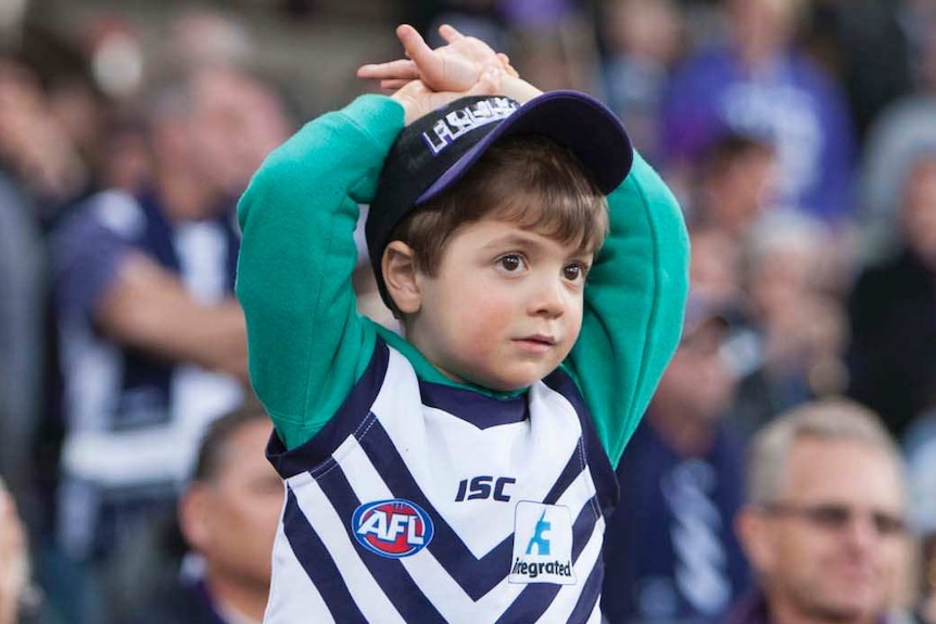 A young boy in a Fremantle guernsey holds his arms over his head amongst the crowd.