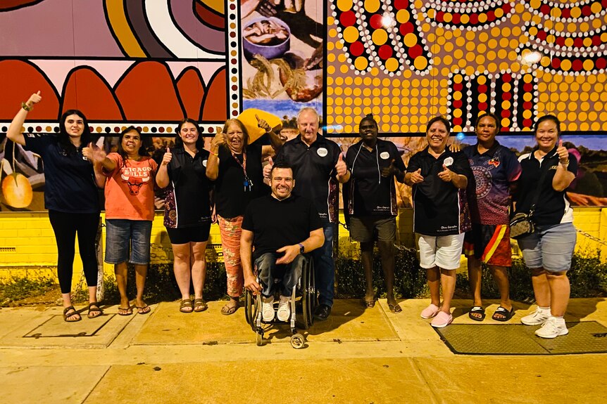 10 people standing in front of an Aboriginal wall mural with thumbs up at night. 