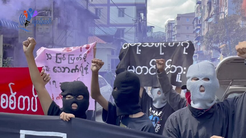 A still image of people in balaclavas protesting with signs in Burmese