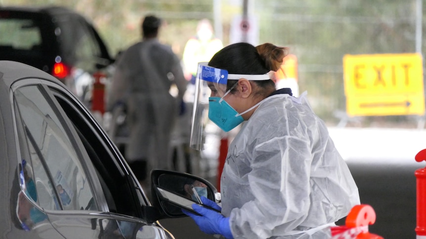 A woman in PPE talks to a driver through a car window.