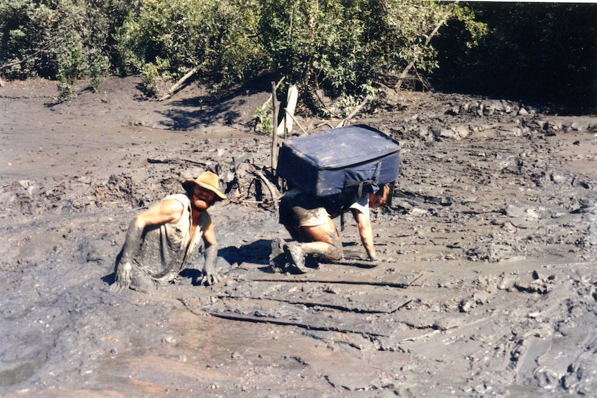 Two men wading through muddy river, one on knees with large suitcase on back.