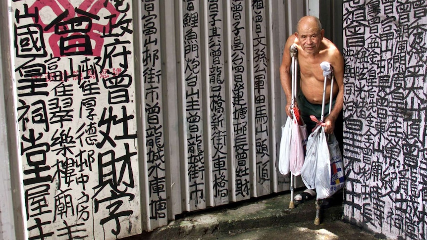 A bare-chested old balding Asian man on crutches holding shopping bags stand against walls graffitied with Chinese characters