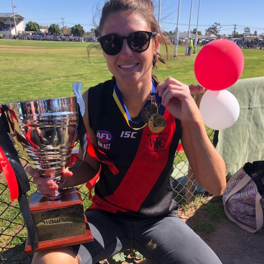 A white woman holding a trophy wearing a black and red afl guernsey smiling. 