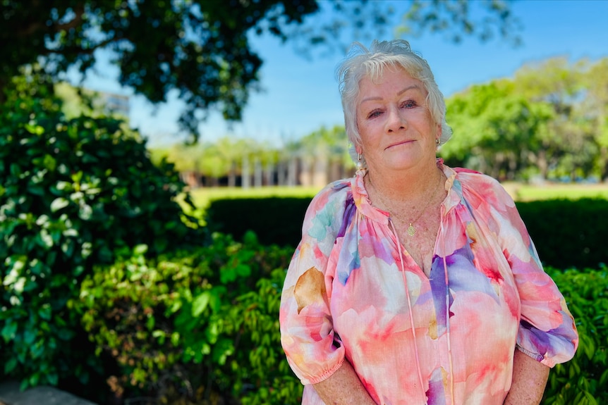An older woman in a colourful top stands in front of some greenery.